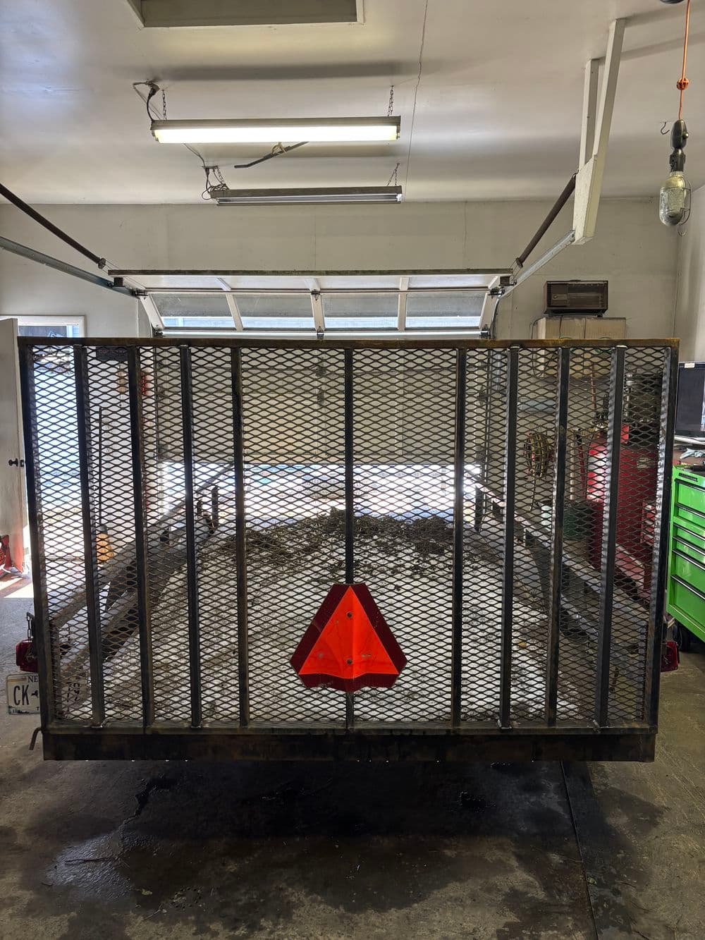 Metal utility trailer with a diamond mesh design and reflective safety triangle in a garage setting.