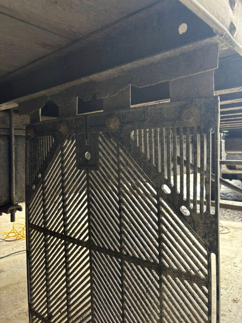 Underside view of a metal grate with diagonal slats, mounted under a heavy structure.